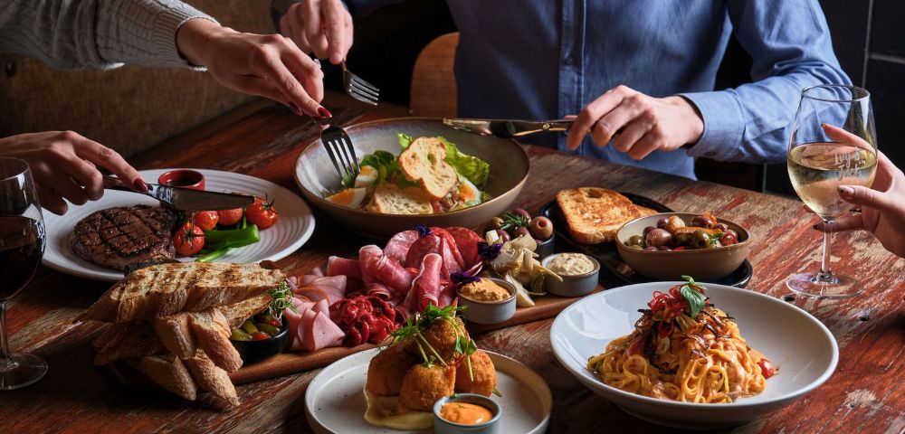 A wooden table with a shared meal featuring steak, salad, pasta, cold cuts, bread, and olives. Three hands reach in, creating a communal, inviting atmosphere.