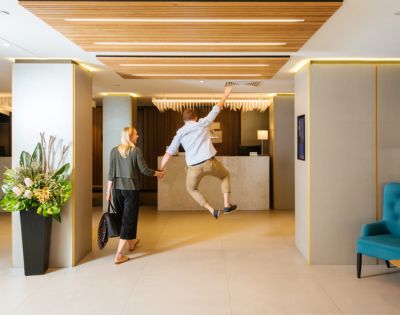 A woman and a man walk hand in hand toward a hotel reception. The man is joyfully leaping in the air. The modern lobby has plants and blue seating.