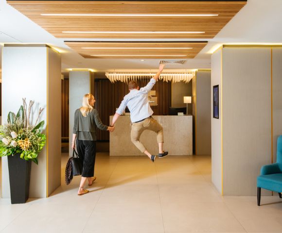 A woman and a man walk hand in hand toward a hotel reception. The man is joyfully leaping in the air. The modern lobby has plants and blue seating.