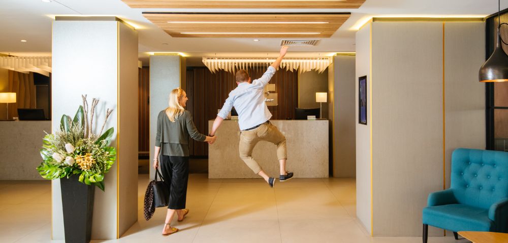 A woman and a man walk hand in hand toward a hotel reception. The man is joyfully leaping in the air. The modern lobby has plants and blue seating.