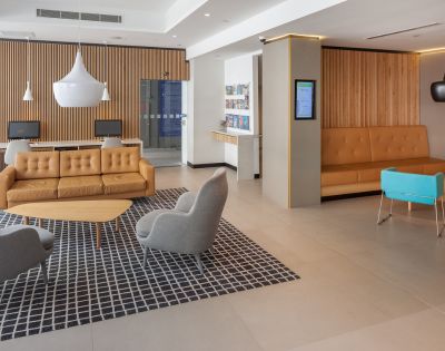 Modern lobby with a warm tone features orange leather sofas, gray chairs, a blue bench, and pendant lights. A checkered rug adds contrast.