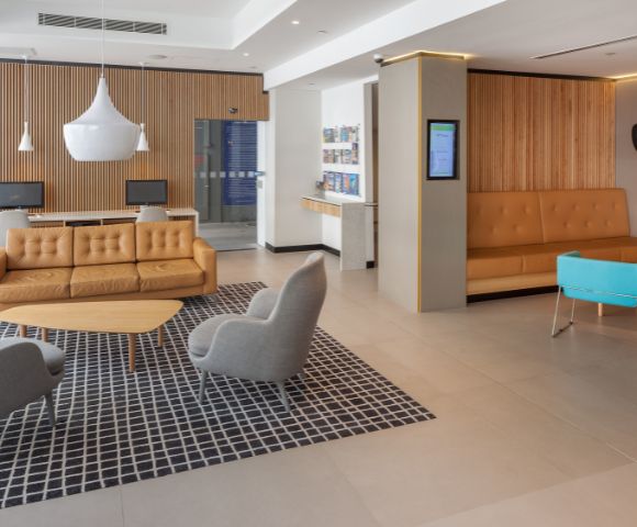 Modern lobby with a warm tone features orange leather sofas, gray chairs, a blue bench, and pendant lights. A checkered rug adds contrast.