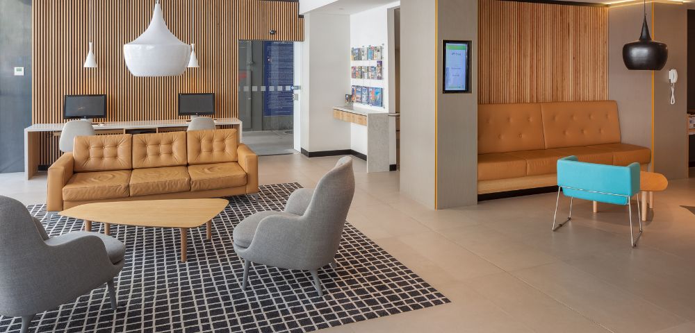 Modern lobby with a warm tone features orange leather sofas, gray chairs, a blue bench, and pendant lights. A checkered rug adds contrast.