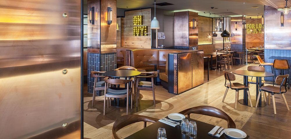 Modern restaurant interior with wooden floors, copper accents, and cozy leather booth seating. Warm ambiance, soft lighting, and neatly set tables.