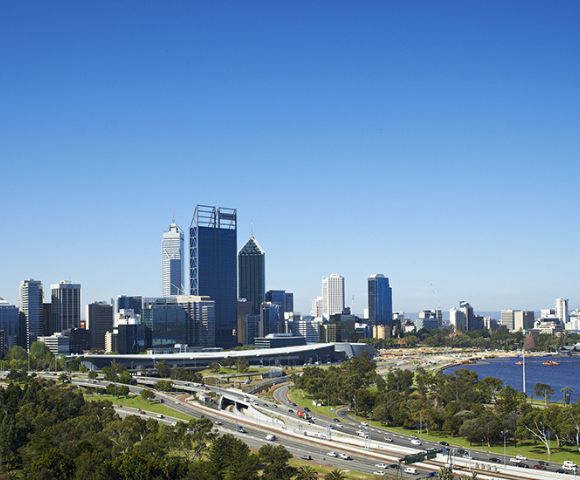 Wide view of a city skyline with tall buildings under a clear blue sky, adjacent to a river. Foreground features lush green parks and a busy highway.