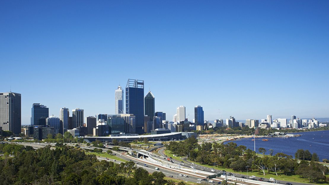 Wide view of a city skyline with tall buildings under a clear blue sky, adjacent to a river. Foreground features lush green parks and a busy highway.