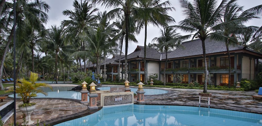 Tropical resort with a serene pool surrounded by tall palm trees and lanterns, reflecting a tranquil ambiance. Two-story buildings are in the background.