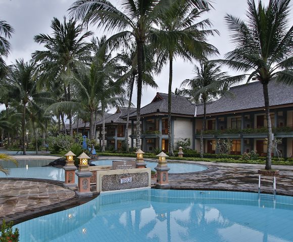 Tropical resort with a serene pool surrounded by tall palm trees and lanterns, reflecting a tranquil ambiance. Two-story buildings are in the background.