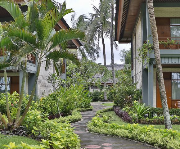 Tropical courtyard with lush greenery, palm trees, and a winding path between two modern buildings. Relaxing atmosphere hints at a resort setting.