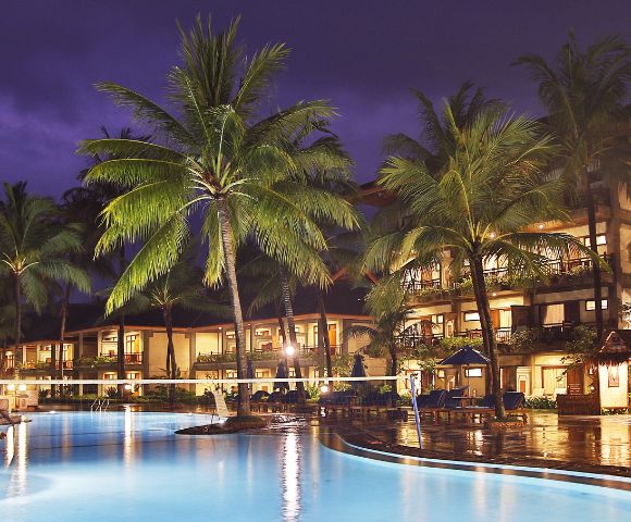 Night scene of a tropical resort with illuminated buildings reflecting in a tranquil pool. Palm trees and lounge chairs create a serene, luxurious atmosphere.