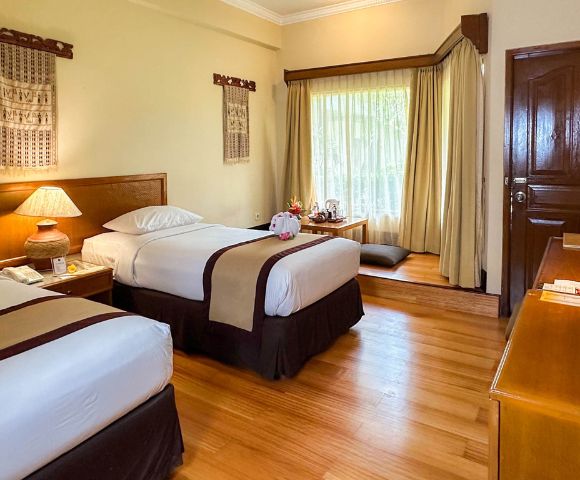 Cozy hotel room with two neatly made twin beds, a wooden lamp, decor with hanging textiles, and a sunlit window nook with cushions, creating a warm, inviting atmosphere.