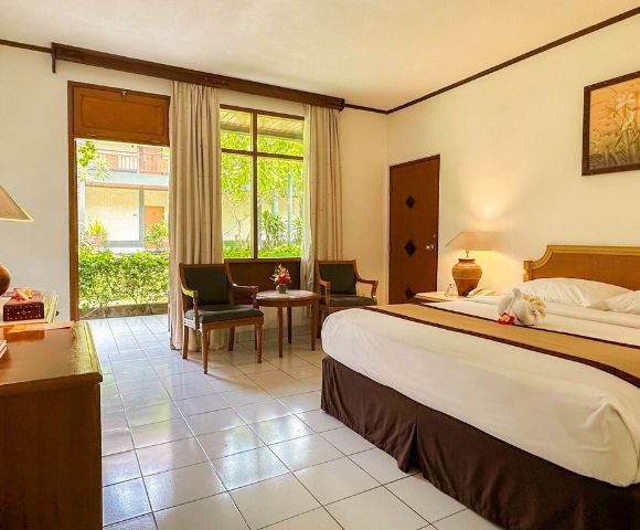 A bright hotel room with a large bed topped with towel swans facing a window with garden views. Two chairs and a table add a cozy touch.