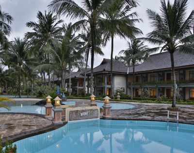 Tropical resort with a serene pool surrounded by tall palm trees and lanterns, reflecting a tranquil ambiance. Two-story buildings are in the background.