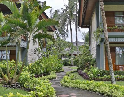 Tropical courtyard with lush greenery, palm trees, and a winding path between two modern buildings. Relaxing atmosphere hints at a resort setting.