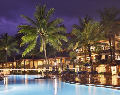 Night scene of a tropical resort with illuminated buildings reflecting in a tranquil pool. Palm trees and lounge chairs create a serene, luxurious atmosphere.