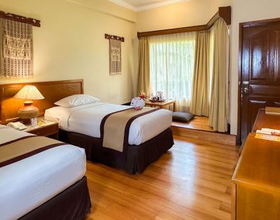 Cozy hotel room with two neatly made twin beds, a wooden lamp, decor with hanging textiles, and a sunlit window nook with cushions, creating a warm, inviting atmosphere.