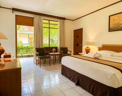 A bright hotel room with a large bed topped with towel swans facing a window with garden views. Two chairs and a table add a cozy touch.