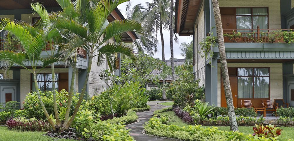 Tropical courtyard with lush greenery, palm trees, and a winding path between two modern buildings. Relaxing atmosphere hints at a resort setting.