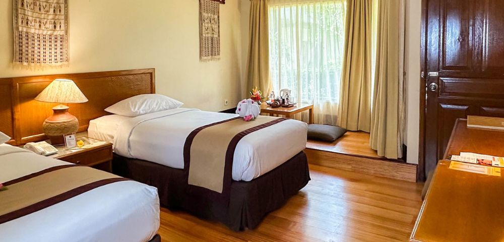 Cozy hotel room with two neatly made twin beds, a wooden lamp, decor with hanging textiles, and a sunlit window nook with cushions, creating a warm, inviting atmosphere.