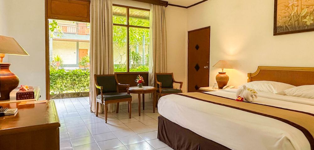 A bright hotel room with a large bed topped with towel swans facing a window with garden views. Two chairs and a table add a cozy touch.