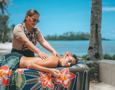 A woman receiving a massage on a table draped with tropical patterned fabric, administered by a smiling masseuse outdoors with ocean and palm trees in the background.