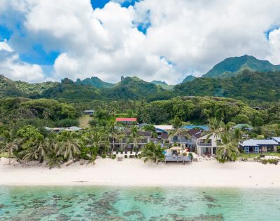 Tropical beach scene with white sand and turquoise water in the foreground. Palm trees and beachfront houses are set against lush, green mountains under a partly cloudy sky.
