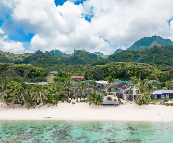 Tropical beach scene with white sand and turquoise water in the foreground. Palm trees and beachfront houses are set against lush, green mountains under a partly cloudy sky.