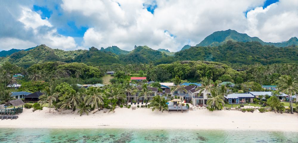 Tropical beach scene with white sand and turquoise water in the foreground. Palm trees and beachfront houses are set against lush, green mountains under a partly cloudy sky.