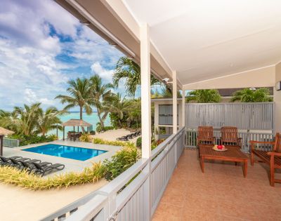 Covered patio with wooden seating overlooks a tropical pool area. Vibrant palm trees and ocean visible under a partly cloudy sky, serene atmosphere.