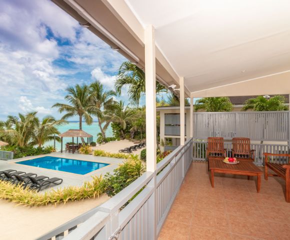 Covered patio with wooden seating overlooks a tropical pool area. Vibrant palm trees and ocean visible under a partly cloudy sky, serene atmosphere.