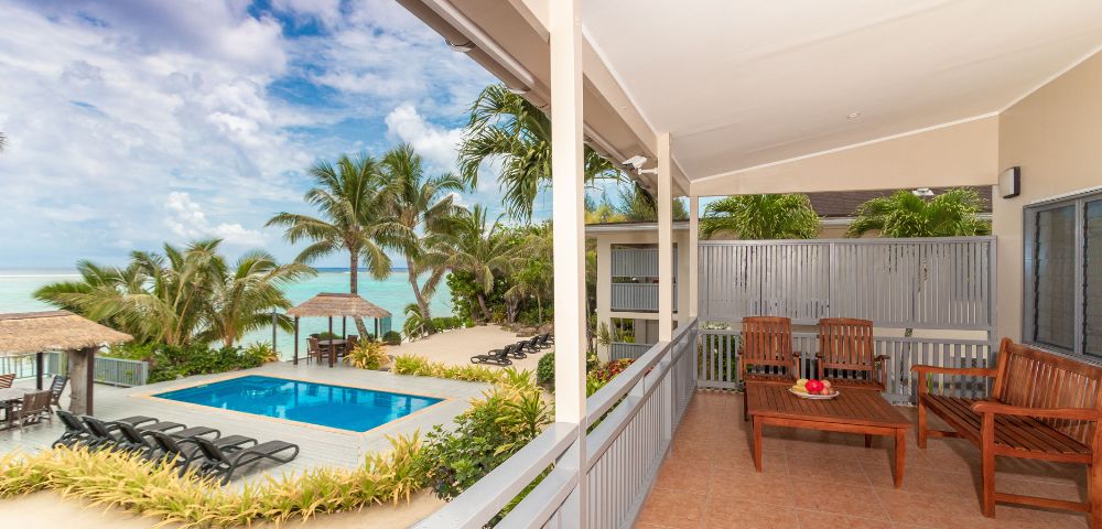 Covered patio with wooden seating overlooks a tropical pool area. Vibrant palm trees and ocean visible under a partly cloudy sky, serene atmosphere.