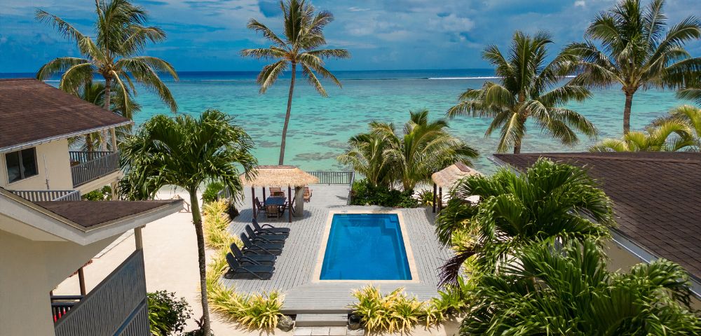 A serene beach resort scene featuring a rectangular pool surrounded by lush palm trees, overlooking a turquoise ocean under a partly cloudy sky.