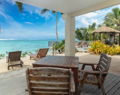 Beachfront patio view with wooden chairs and tables, overlooking turquoise ocean under a clear blue sky. Palm trees and thatched huts add a tropical feel.