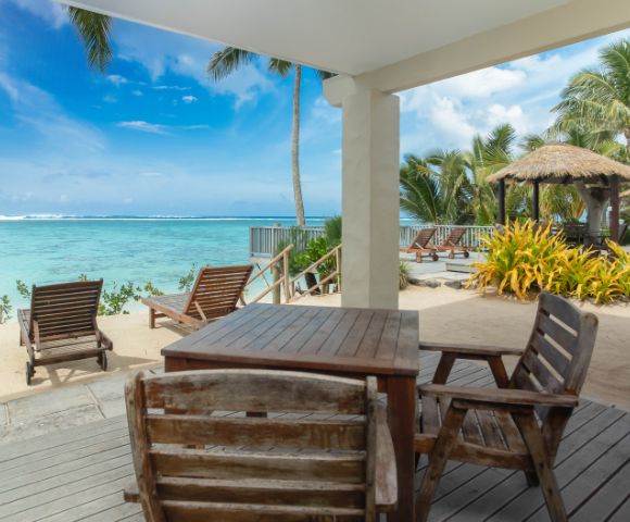 Beachfront patio view with wooden chairs and tables, overlooking turquoise ocean under a clear blue sky. Palm trees and thatched huts add a tropical feel.