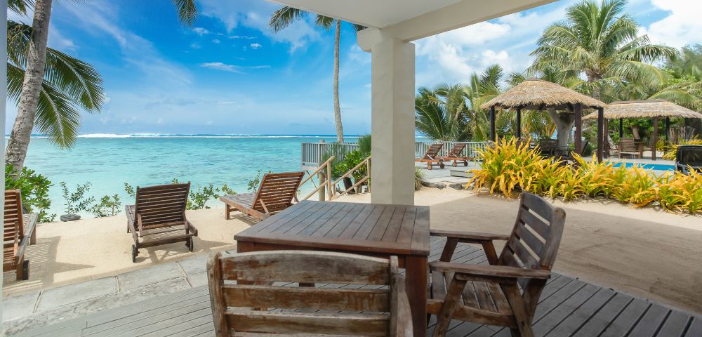 Beachfront patio view with wooden chairs and tables, overlooking turquoise ocean under a clear blue sky. Palm trees and thatched huts add a tropical feel.