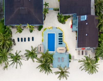 Aerial view of a beachfront property with a pool surrounded by palm trees. Clear turquoise water and sandy shore create a tranquil, tropical vibe.