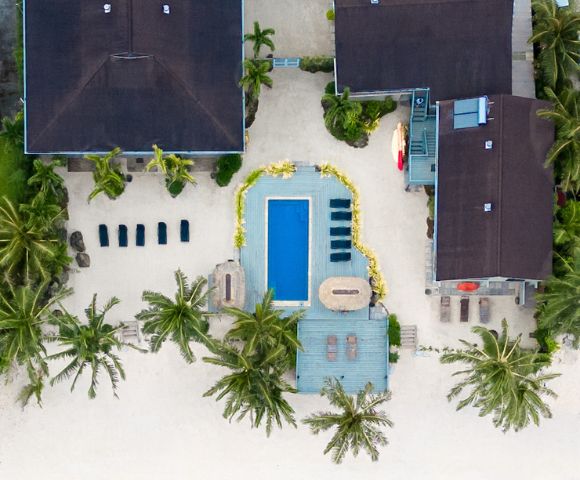 Aerial view of a beachfront property with a pool surrounded by palm trees. Clear turquoise water and sandy shore create a tranquil, tropical vibe.