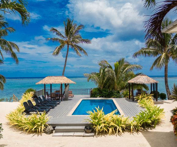 Tropical beach scene with a rectangular pool, surrounded by lounge chairs and lush greenery. Thatched huts and palm trees overlook the blue ocean.