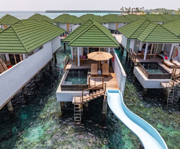 Overwater bungalows with green roofs, featuring private decks, pools, and a waterslide into the clear ocean below. Relaxing, tropical getaway vibe.