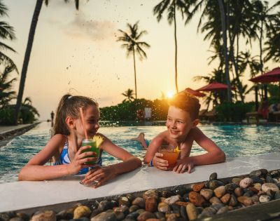 Two smiling children enjoy drinks on a pool's edge at sunset, surrounded by palm trees and red umbrellas, creating a joyful, tropical atmosphere.