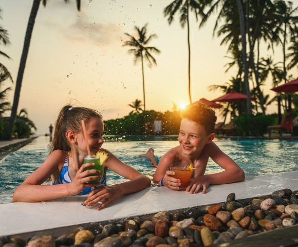 Two smiling children enjoy drinks on a pool's edge at sunset, surrounded by palm trees and red umbrellas, creating a joyful, tropical atmosphere.