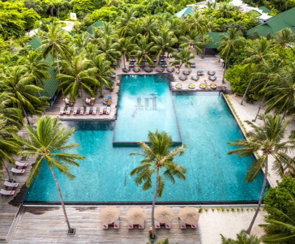 Aerial view of a tropical resort with two large, blue swimming pools surrounded by lush palm trees. Lounge chairs and umbrellas line the pool deck. Relaxing atmosphere.