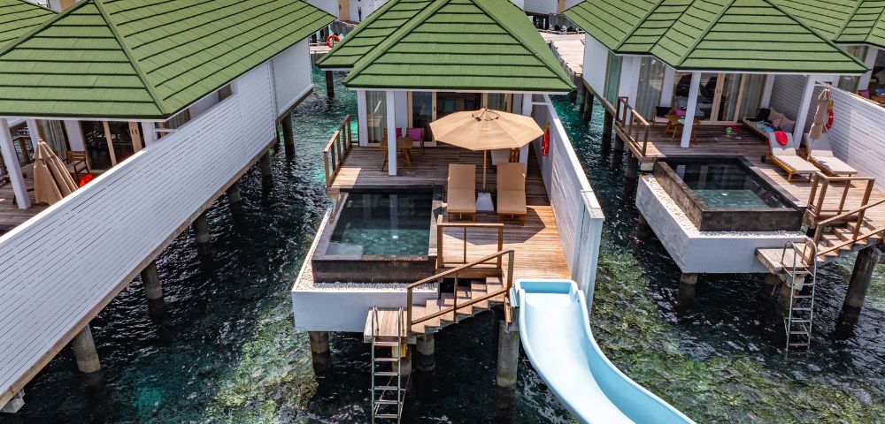 Overwater bungalows with green roofs, featuring private decks, pools, and a waterslide into the clear ocean below. Relaxing, tropical getaway vibe.