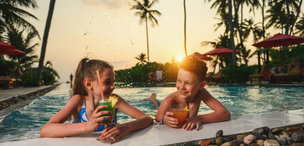 Two smiling children enjoy drinks on a pool's edge at sunset, surrounded by palm trees and red umbrellas, creating a joyful, tropical atmosphere.