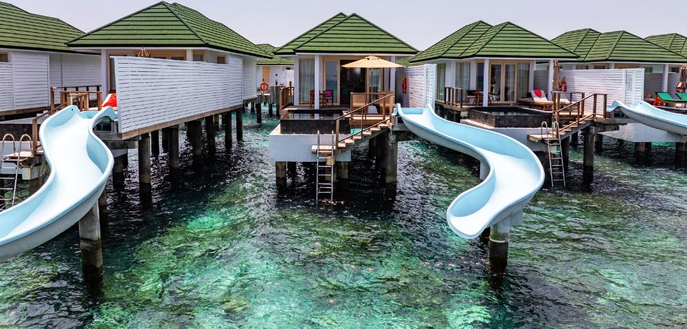 Overwater bungalows with green roofs feature spiral slides leading into the clear turquoise ocean, creating a playful and serene tropical scene.
