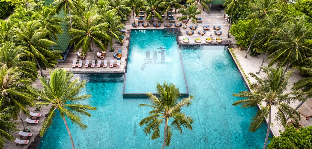 Aerial view of a tropical resort with two large, blue swimming pools surrounded by lush palm trees. Lounge chairs and umbrellas line the pool deck. Relaxing atmosphere.