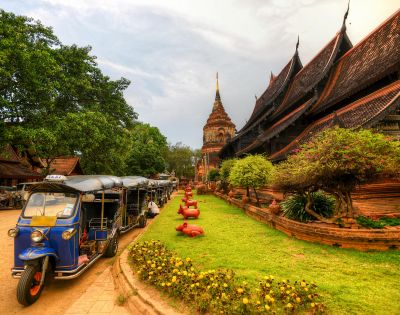 Ancient temple with steeply sloped roofs beside lush greenery and a blue tuk-tuk. The scene is vibrant, peaceful, with a touch of cultural charm.