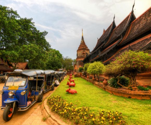 Ancient temple with steeply sloped roofs beside lush greenery and a blue tuk-tuk. The scene is vibrant, peaceful, with a touch of cultural charm.
