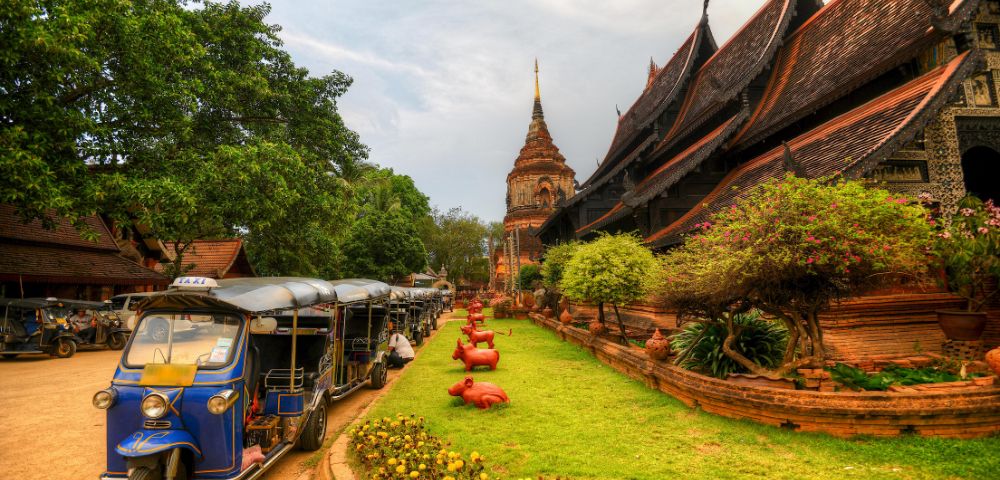 Ancient temple with steeply sloped roofs beside lush greenery and a blue tuk-tuk. The scene is vibrant, peaceful, with a touch of cultural charm.