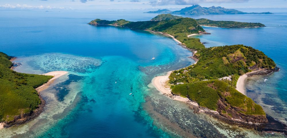 Aerial view of lush green islands surrounded by vibrant blue waters and coral reefs, with a sandy beach and distant mountains.